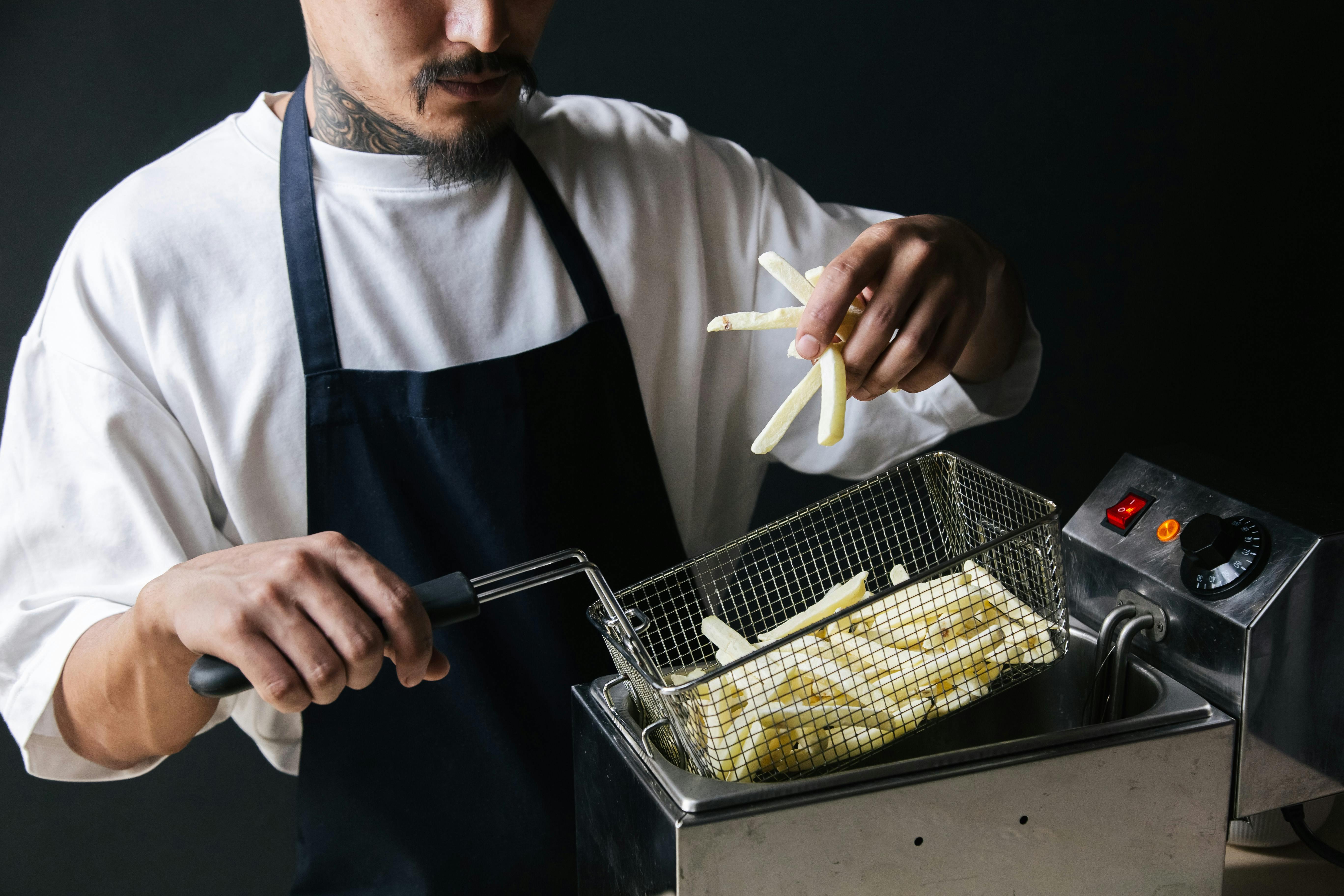A chef handling raw french fries with a strainer basket in a deep fryer.